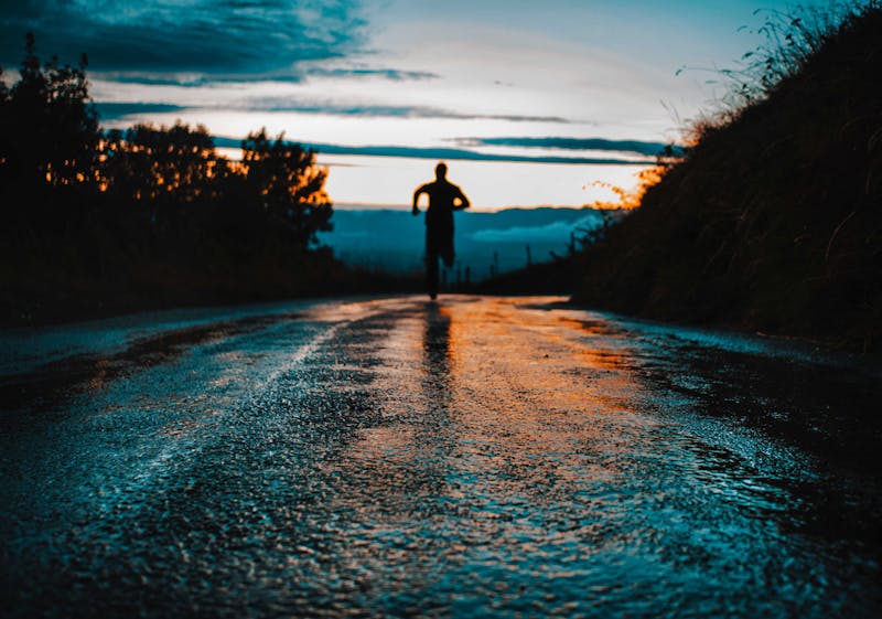 A man running on a road into the distance. The road is wet and his figure is a silhouette.