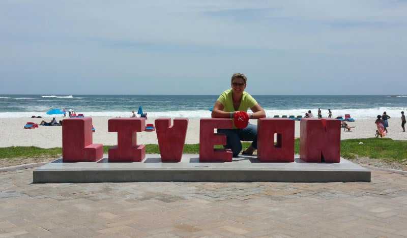 Anita on camps bay beach crouching behind big letters: "LIVE ON".