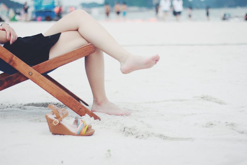 a woman sitting on a beach on a wooden chair with her legs crossed with one shoe in sight.