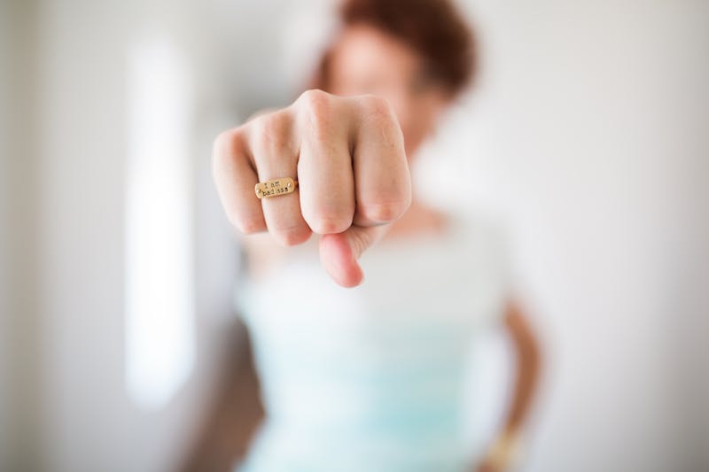 I woman standing with a white jersey on holding her fist out in front of her face there is a ring on her finger on her saying bad ass