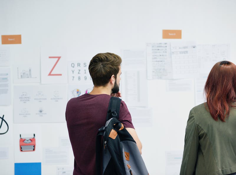 Two co-workers looking at a wall of printed documents on a wall. Shot is taken from behind. One of the two has a bag over the one shoulder.