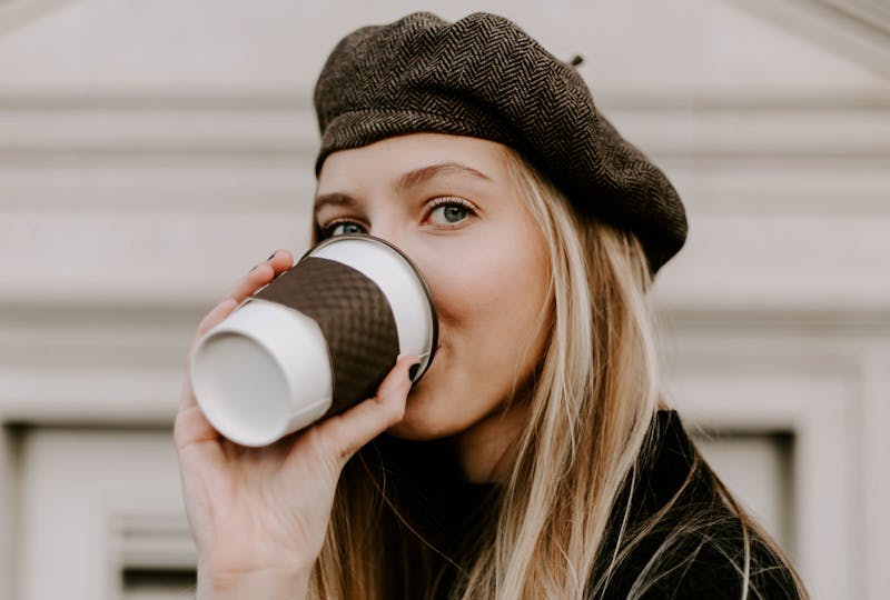 A blond woman with a black woolen beanie on drinking coffee out of a take away cup