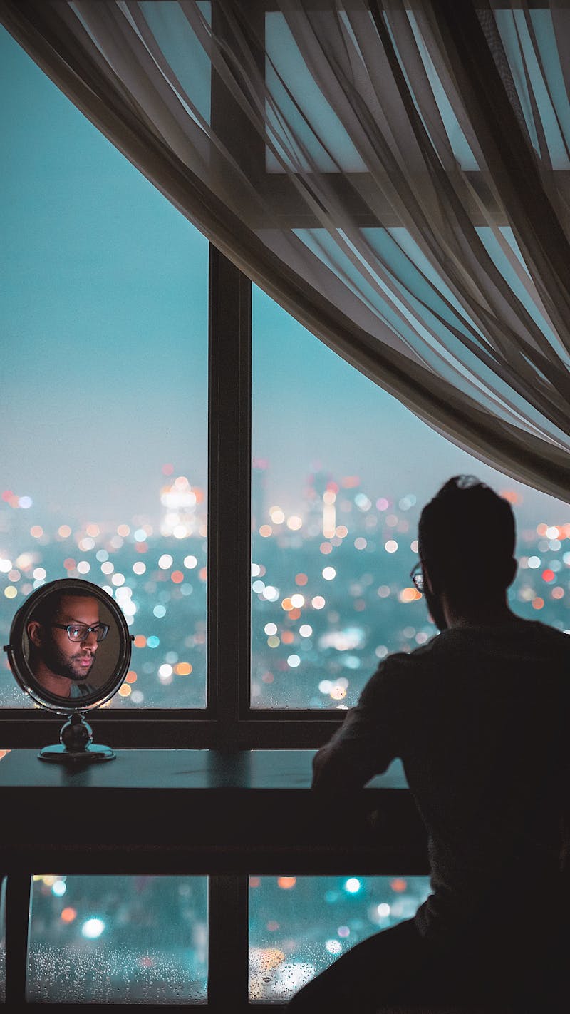 A man with a beard with his back to us looking into a mirror while looking at a view from an apartment overlooking the city