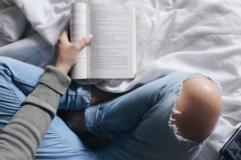 A girl sitting cross-legged on a bed with ripped jeans on and a book on the bed open