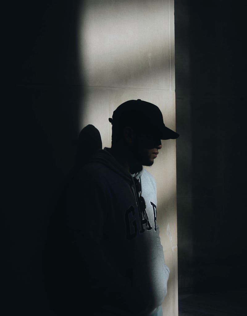 A guy standing in the shadows; wearing a cap; wearing a GAP hoody.