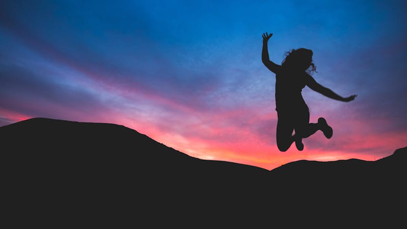 A girl jumping, joyously. Her figure is a silhouette. In the background, there is a sunset and a mountain.