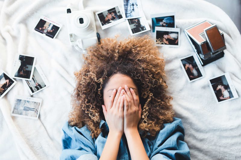 A girl laying on a bed with her hands over her eyes with Polaroid pictures around her.