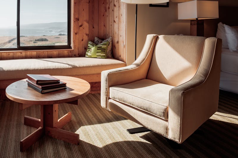 A single beige chair at a window looking out at the ocean. Books on the coffee table