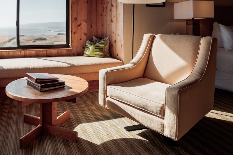 A single beige chair at a window looking out at the ocean.  Books on the coffee table