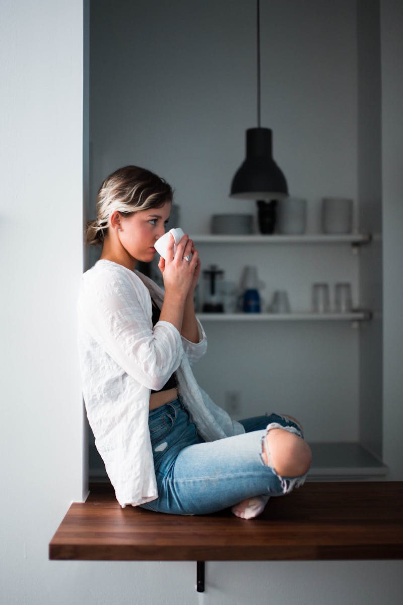 A blond girl wearing a white jersey, sitting on a ledge holding a coffee cup to her mouth with both hands.