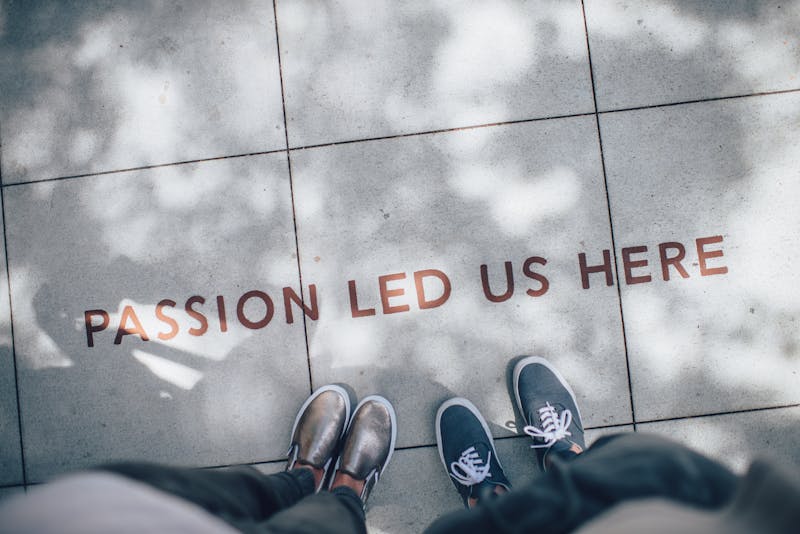 Text on concrete that reads "PASSION LED US HERE". Two people's feet are pointed toward the text.