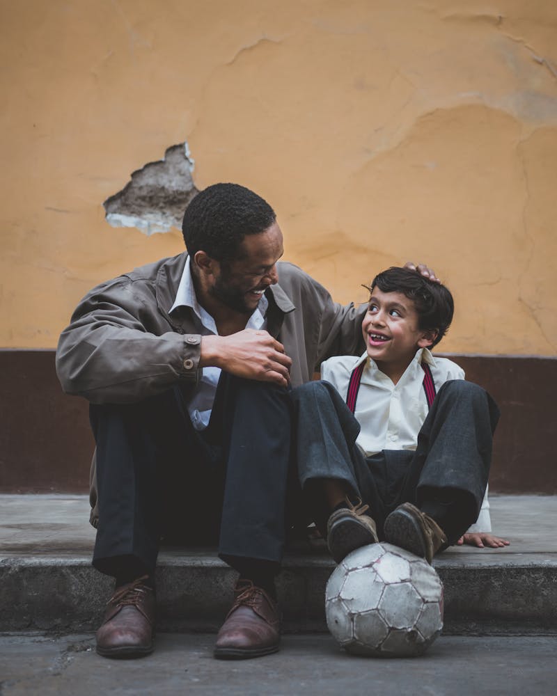 Man sitting with a young boy on the payment. Little boy smiling and holding a soccer ball under his feet