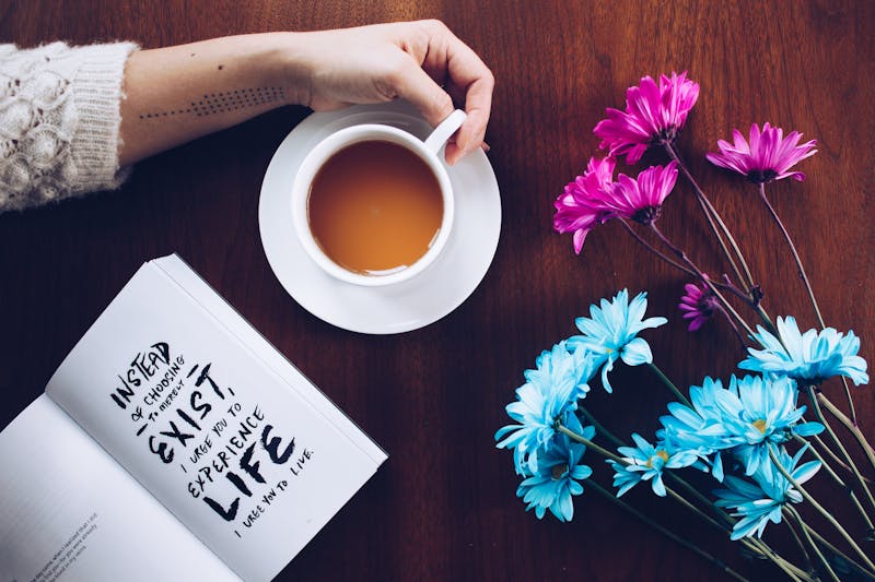 A womans arm touching a coffee mug with a book lying next to it with some blue flowers on a desk