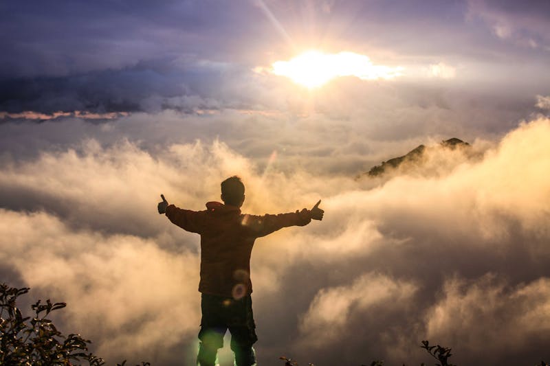 A man who seems to be standing at the top of a mountain gesturing with 2x thumbs up.